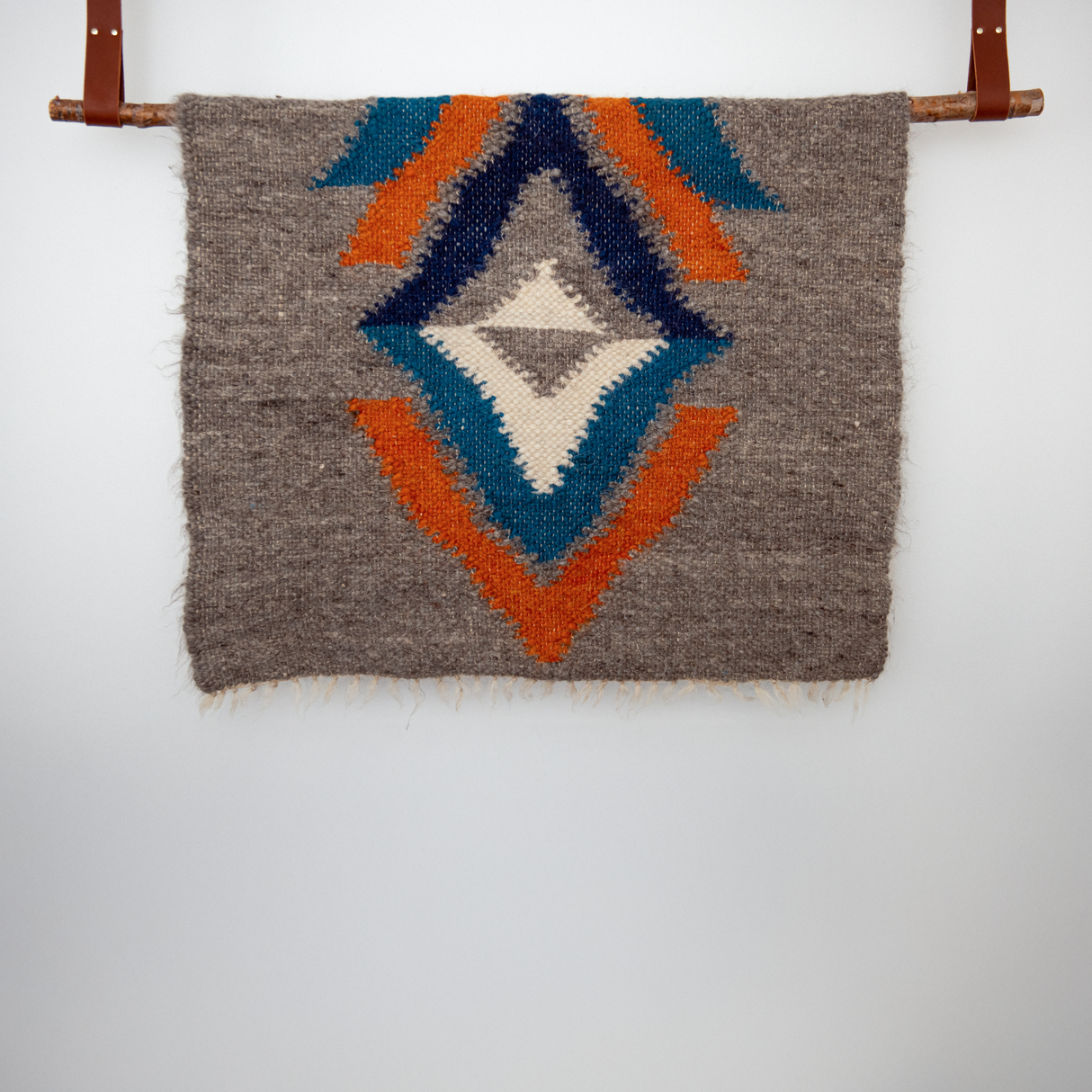 The Podyh (Breathe) Wool Statement Throw / Baby Blanket, featuring geometric patterns in blue, orange, and cream, hangs from a wooden rod on the wall.