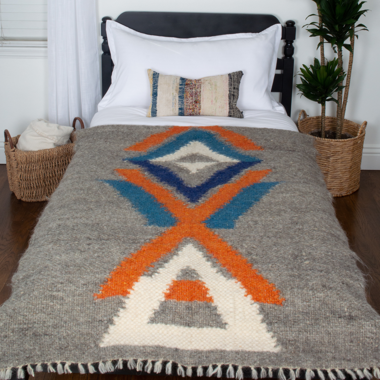 A bed with white pillows, baskets on a wooden floor, and the Podyh (Breathe) Wool Statement Throw / Baby Blanket in gray, orange, and blue tribal patterns.