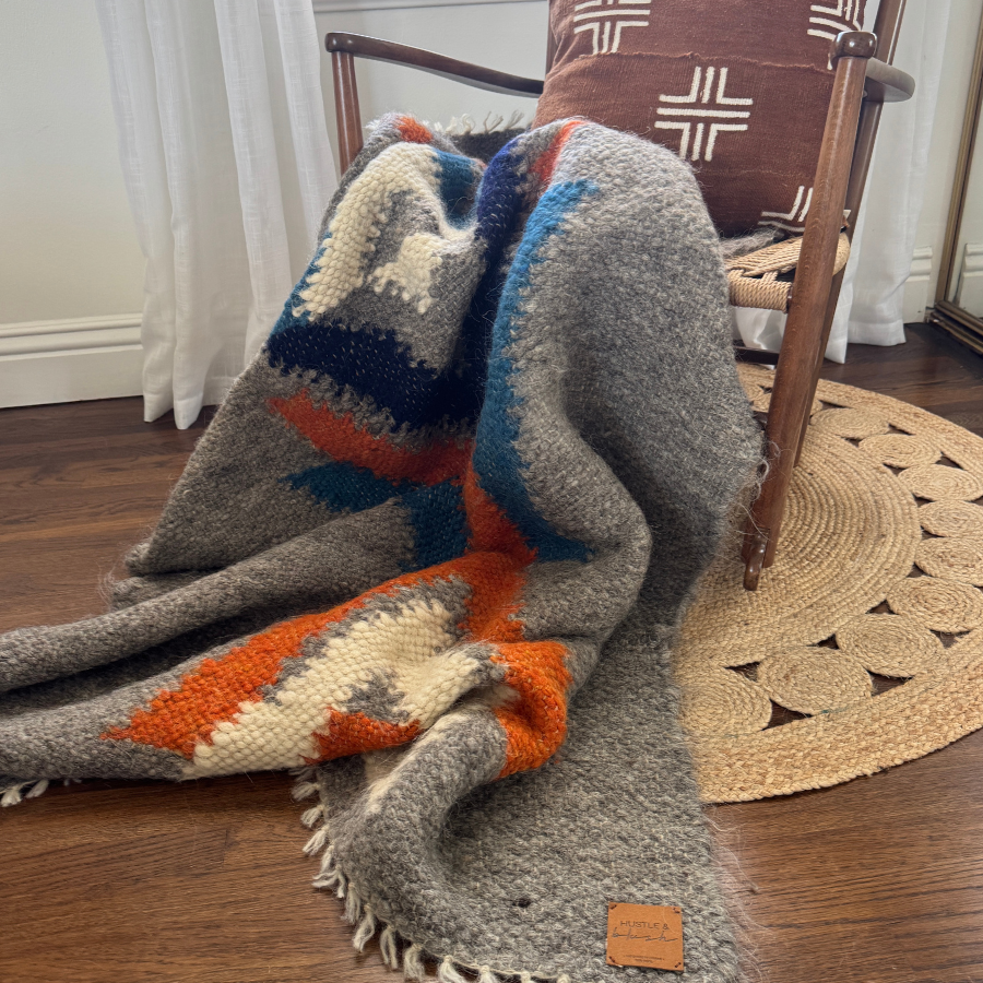 The Podyh (Breathe) Wool Statement Throw is draped over a wooden chair with a geometric pillow, set atop a wooden floor and round rug.