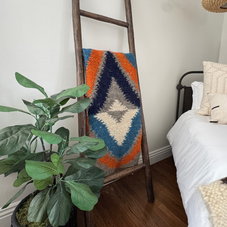 The Podyh (Breathe) Wool Statement Throw / Baby Blanket, handcrafted in vibrant colors, hangs on a wooden ladder next to a bed and potted plant, adding warmth to a cozy room.