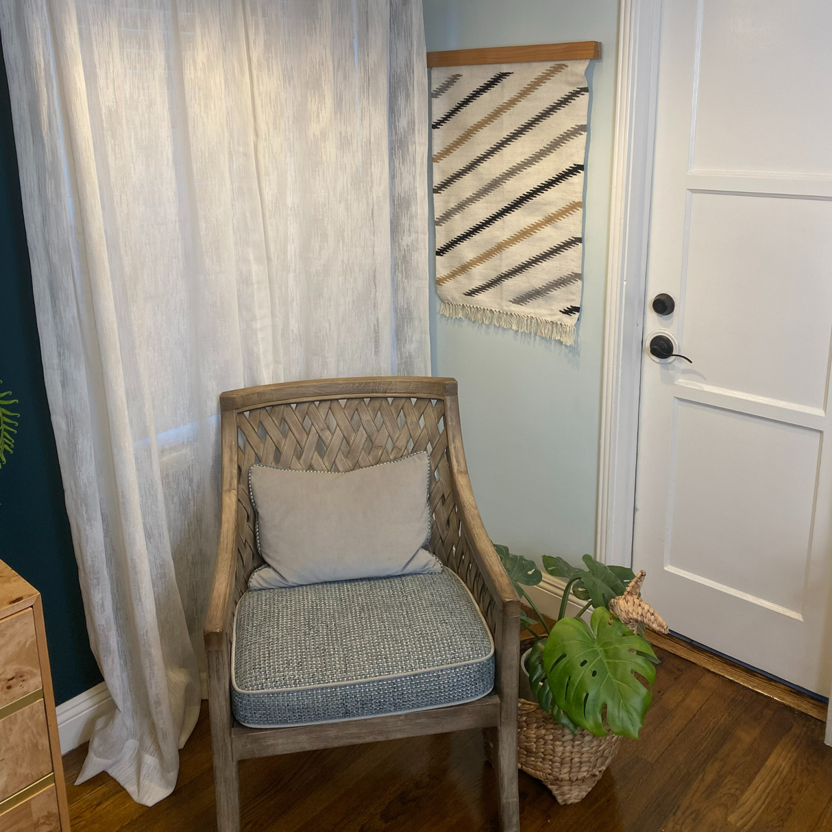 A Promin (Beam) Wool Rug / Wall Hanging, handwoven wicker chair with blue cushion, plant, and sheer curtains create a cozy nook by a white door—perfect for relaxing.