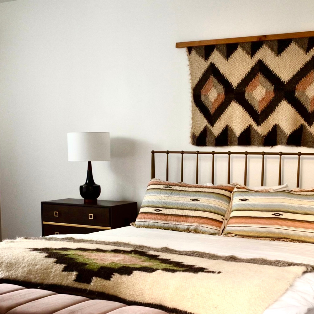 Minimalist bedroom with a metal bed, striped bedding, and a lamp on a nightstand, featuring statement wall decor: the Svitlo (Bright) Wool Wall Hanging.