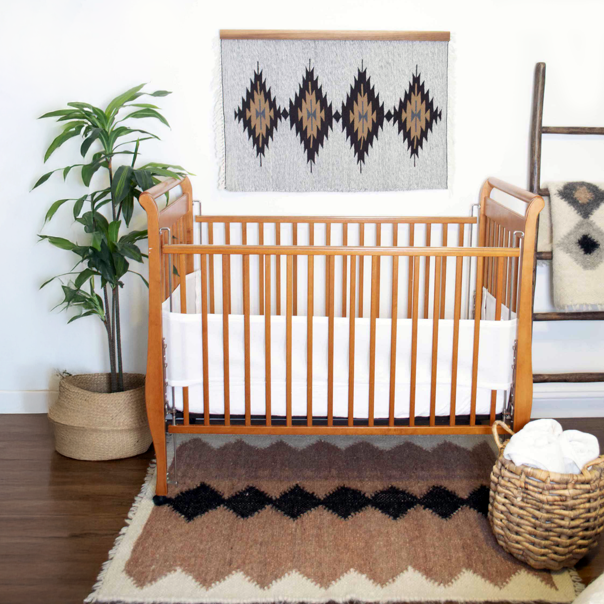 A wooden crib sits in a cozy nursery with plants, geometric wall art, and the Smilyv (Bold) Wool Rug / Wall Hanging, adding warmth and subtle elegance with its neutral tones.