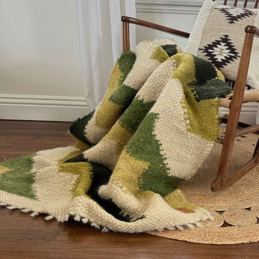 The Sontse (Sun) Wool Statement Throw / Baby Blanket in green, black, and cream is draped over a wooden rocking chair next to a patterned pillow.