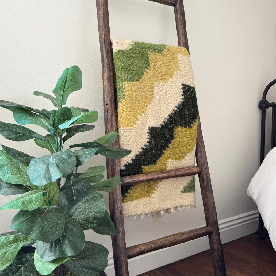 The Sontse (Sun) Wool Statement Throw / Baby Blanket, featuring green, yellow, and cream stripes, hangs on a wooden ladder beside a leafy plant.