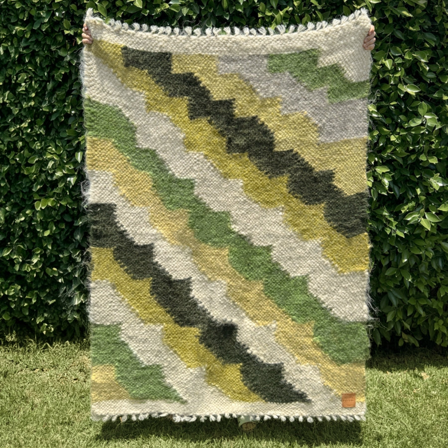 A person holds the Sontse (Sun) Wool Statement Throw / Baby Blanket, featuring a textured green and yellow zigzag pattern, in front of leafy bushes.