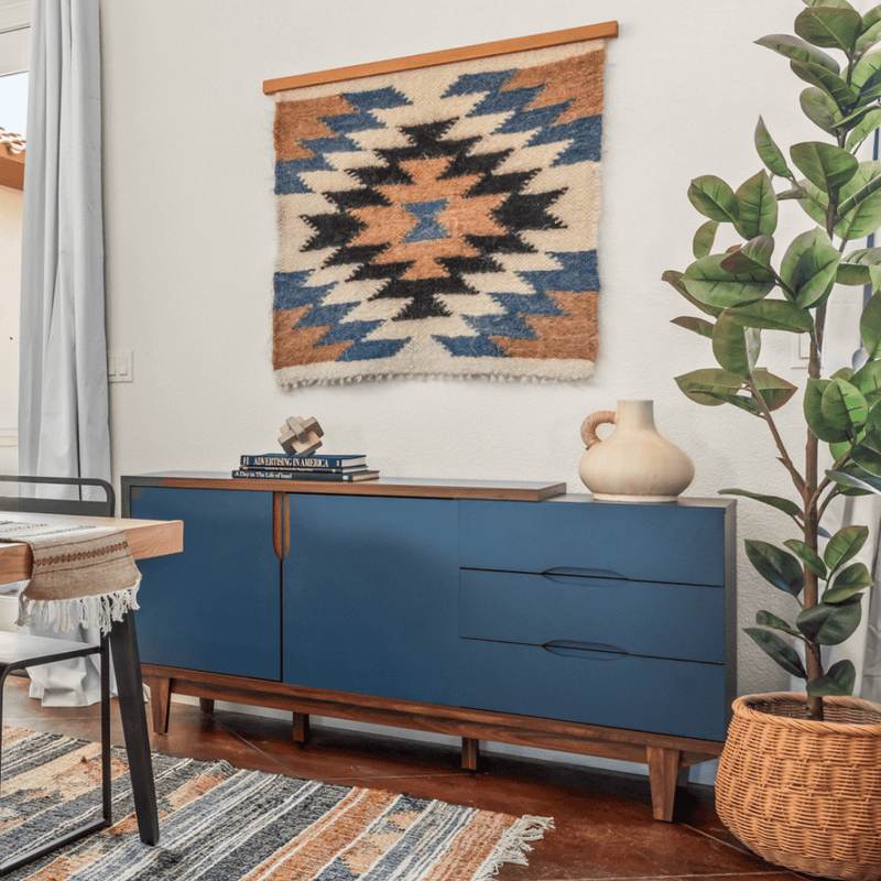 Blue sideboard with books, a vase, and a plant beside it; geometric wall hanging above.