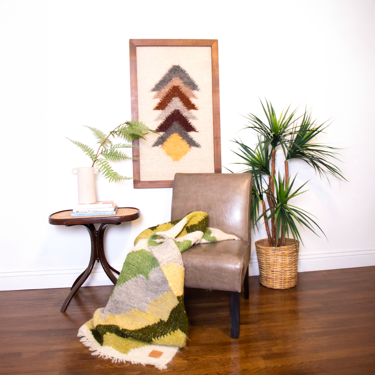 A tan chair with a green blanket sits by plants and a side table, beneath the Strilka (Arrow) Wool Framed Wall Art and geometric accents above.