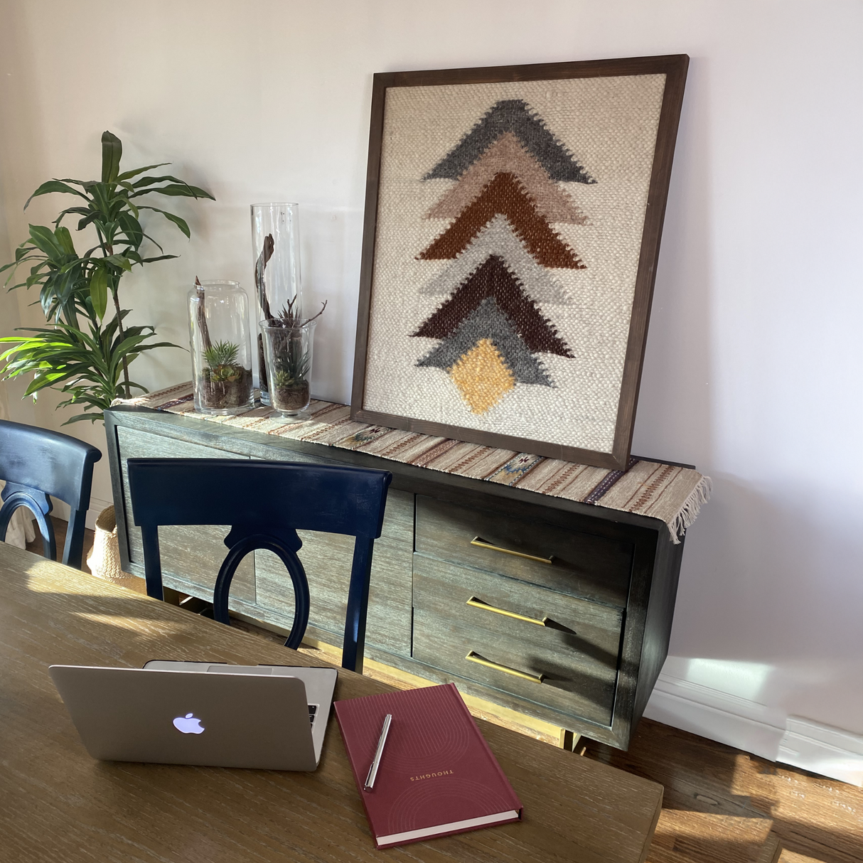 A laptop and notebook sit on a table by a sideboard with plants and Strilka (Arrow) Wool Framed Wall Art, adding eco-friendly geometric accents to the home decor.