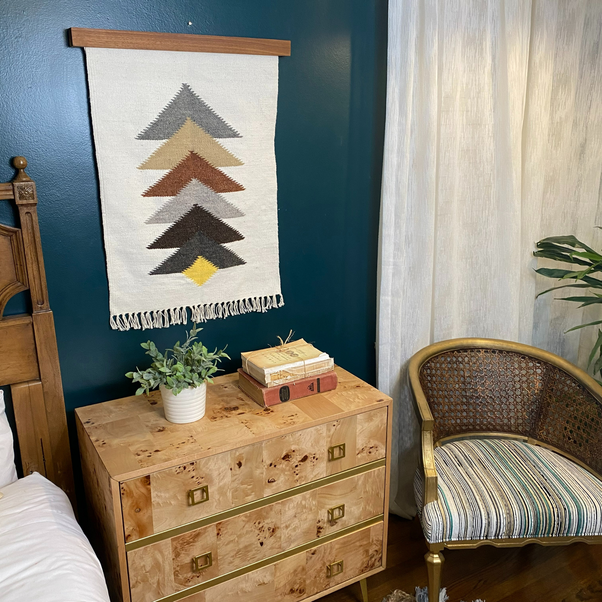 The Strilka (Arrow) Wool Rug / Wall Hanging with chevron V shapes decorates the wall above a dresser with books and a plant, complemented by a nearby gold-accented chair for a vibrant touch.