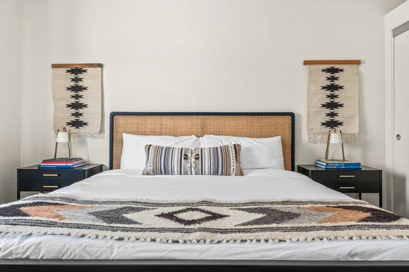 A neatly made bed with patterned pillows and blanket, flanked by two nightstands and wall hangings.