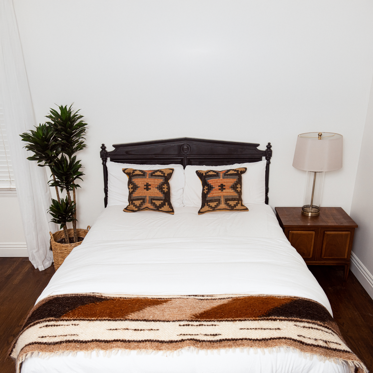 A neatly made bed features the Tak (Yes) Wool Bed / Couch Runner, patterned pillows, a brown blanket, a handcrafted rug, a plant, and a lamp on a wooden nightstand.
