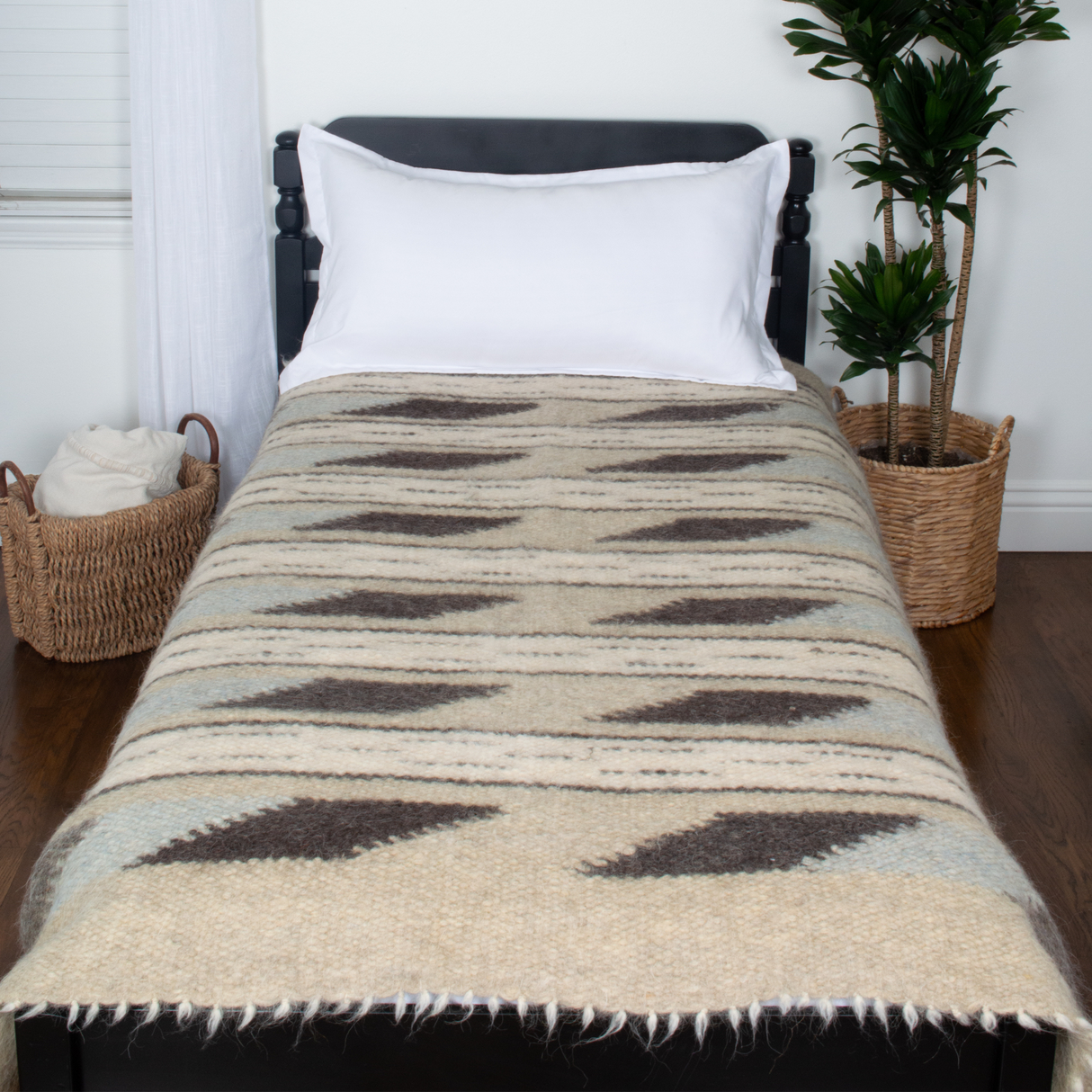 A single bed features the Tak (Yes) Wool Statement Bed Blanket, a white pillow, and baskets beside it, next to a green potted plant.