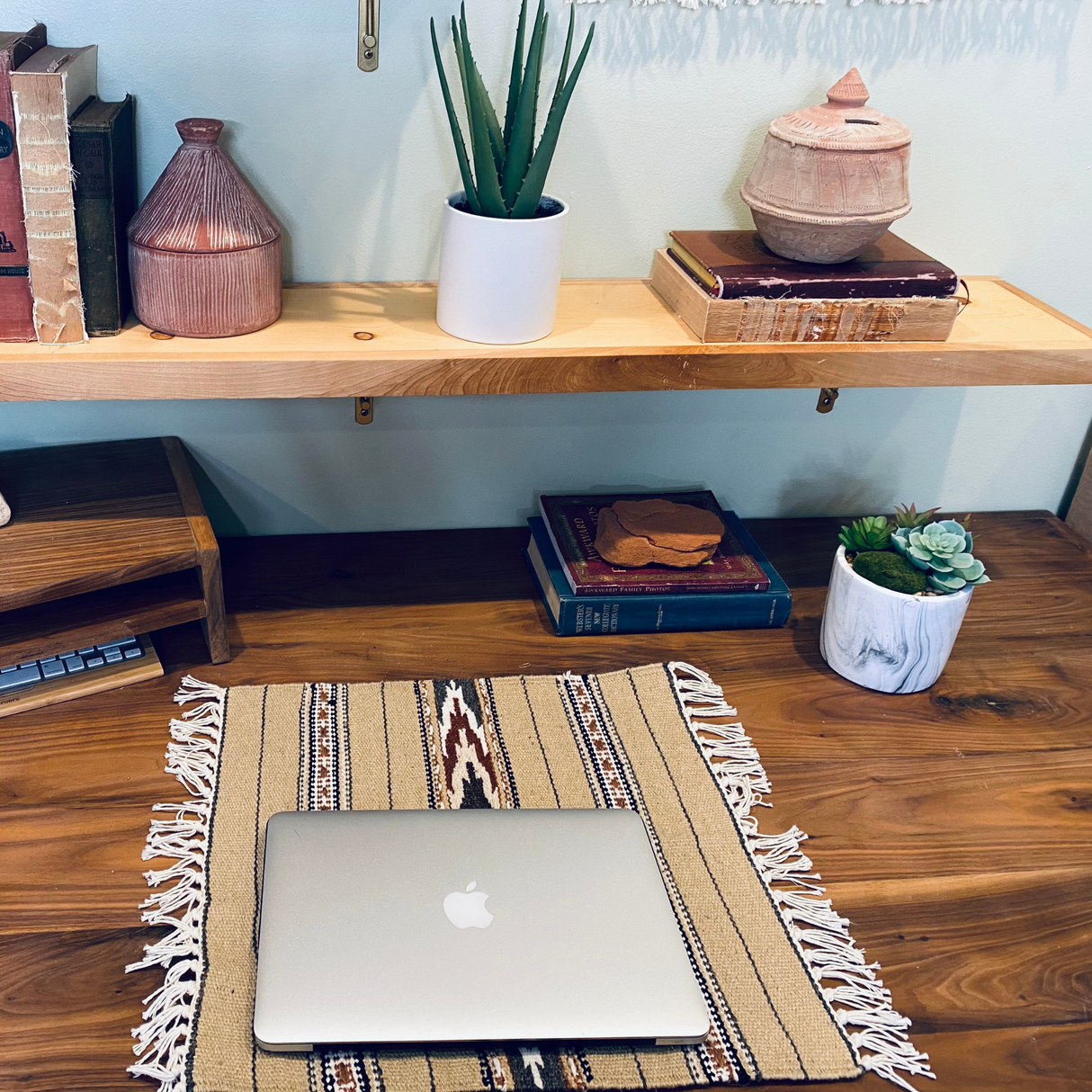 A closed MacBook sits on the Teplota (Warmth) Wool Small Rug, surrounded by books, plants, and pottery—adding cozy style to a wooden desk and shelves.