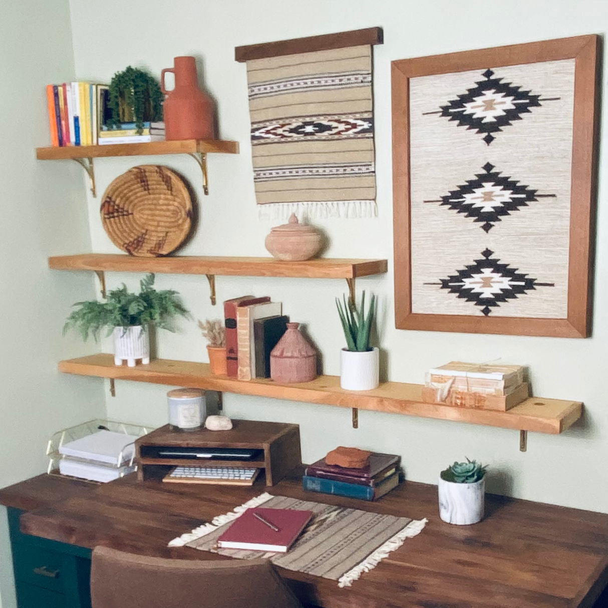 A tidy desk with books, plants, and decor sits beneath three wooden shelves, two woven wall hangings, and a Teplota (Warmth) Wool Small Rug / Mat.
