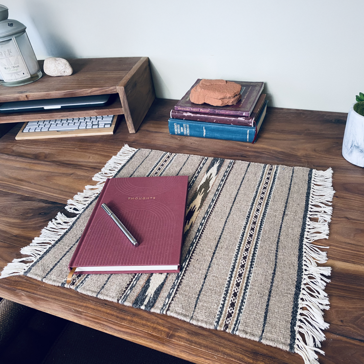 The Teplota (Warmth) Wool Small Rug / Mat - Dark sits on a wooden desk with a journal, pen, books, candle, and plant—ideal desk decor for creating an inviting workspace.