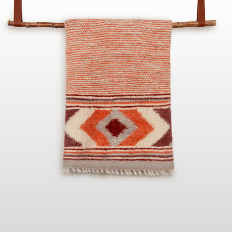The Tkach (Weaver) Wool Statement Bed Blanket, featuring orange and cream geometric patterns and handcrafted from Carpathian sheep wool, hangs gracefully on a wooden rod.