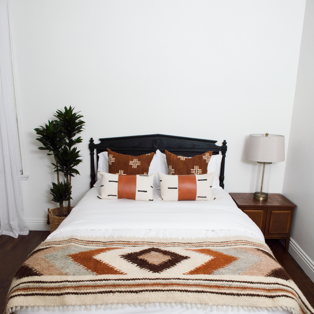 A neatly made bed features patterned pillows and a Tkach (Weaver) Wool Bed/Couch Runner, next to a plant and a lamp on a wooden nightstand.