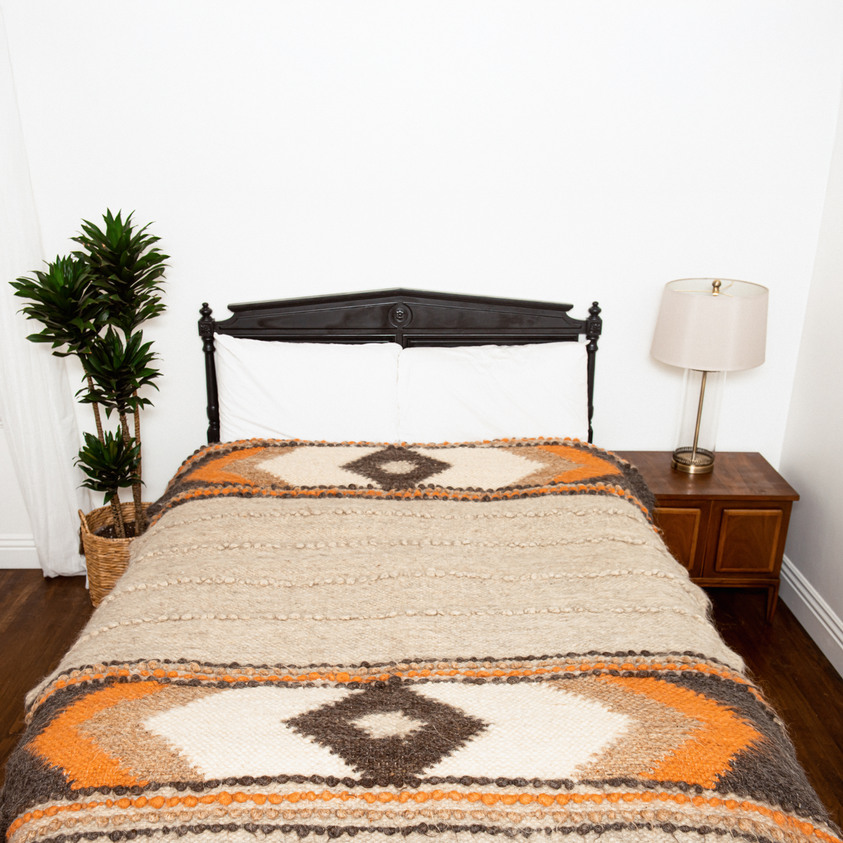 A bed with the Tkach (Weaver) Wool Statement Bed Blanket, a nightstand with a lamp, and a potted plant beside it.