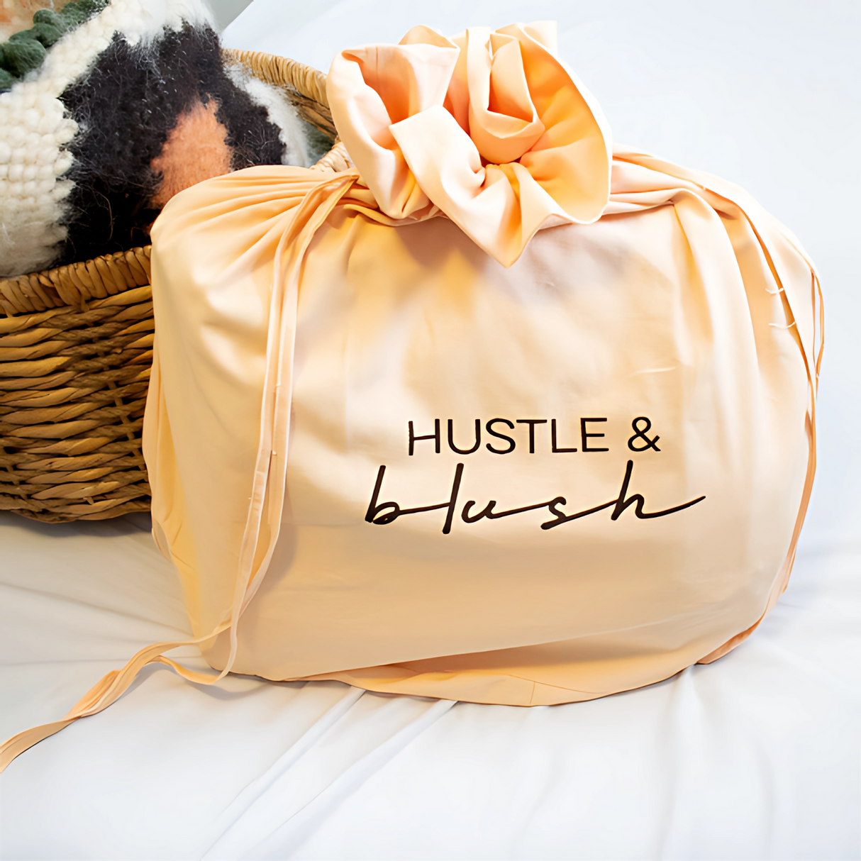 A peach linen laundry bag with "HUSTLE & blush" text sits beside a woven basket and the Tkach (Weaver) Wool Statement Bed Blanket on a white surface.