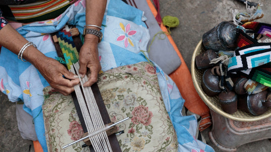 A person weaves colorful textiles by hand while sitting on patterned blankets and cushions.