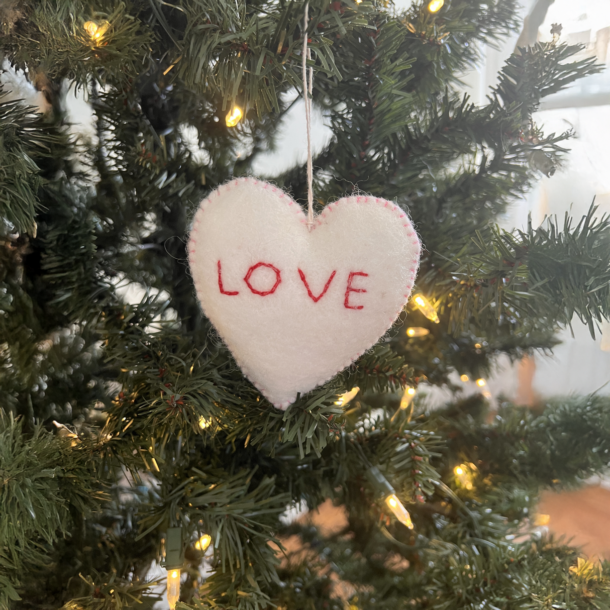 A trio of hand-stitched felt star and heart ornaments, crafted from sustainable wool, hangs on a Christmas tree adorned with white lights.