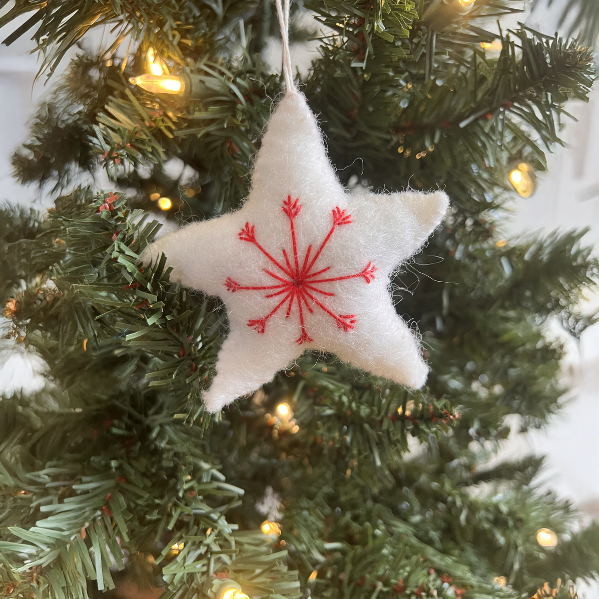 A star-shaped ornament from the Trio of Hand-Stitched Felt Stars and Heart Ornaments collection, crafted from sustainable wool, with red embroidery, hangs on a lit Christmas tree.
