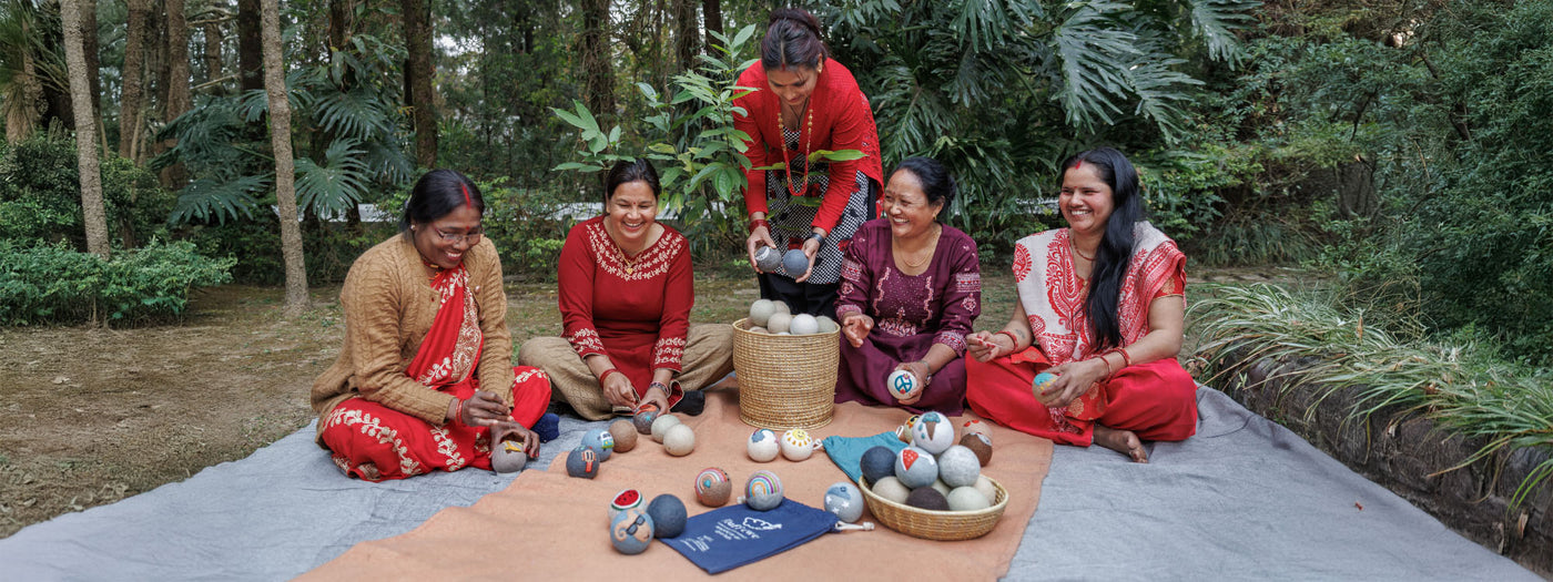 Five women sit outdoors on a mat, smiling and crafting handmade items from natural materials.