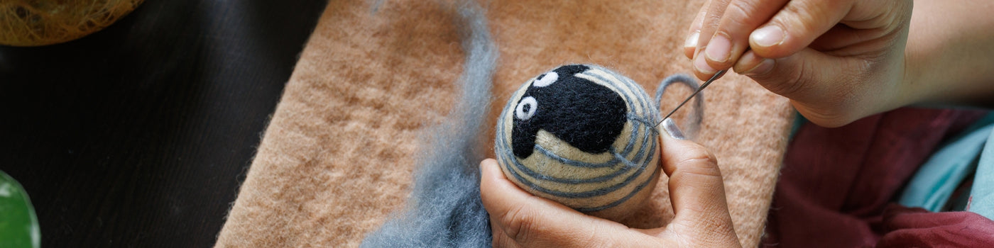 Close-up of hands needle felting a black and white sheep design onto a round wool object with a felting needle.