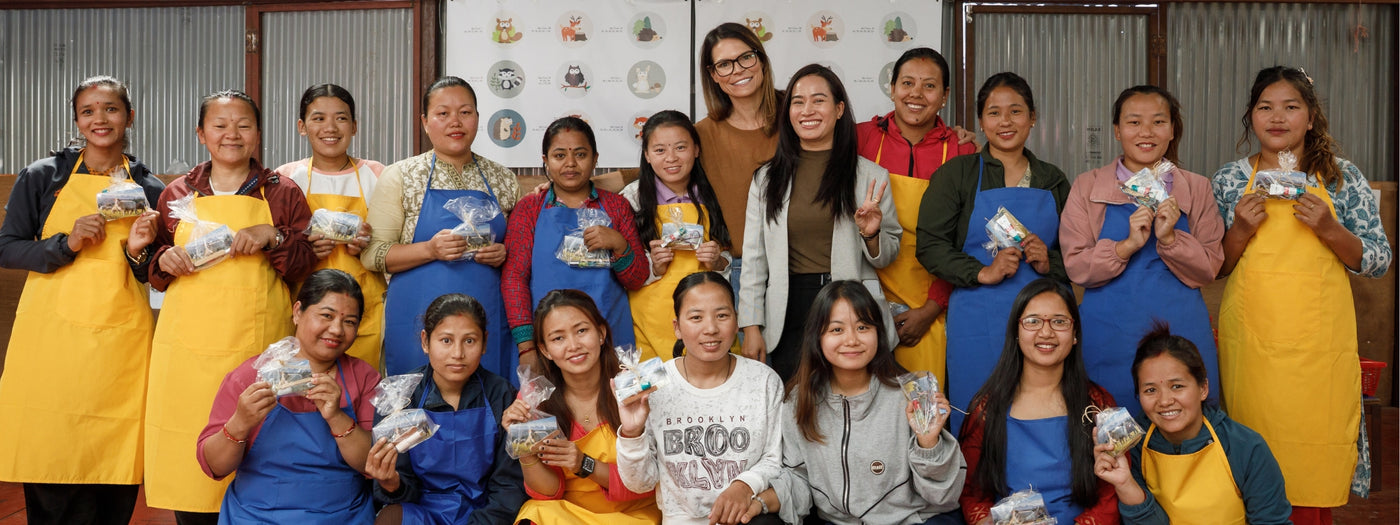 Group of women, some in aprons, smiling and holding wrapped snacks, posing together indoors.