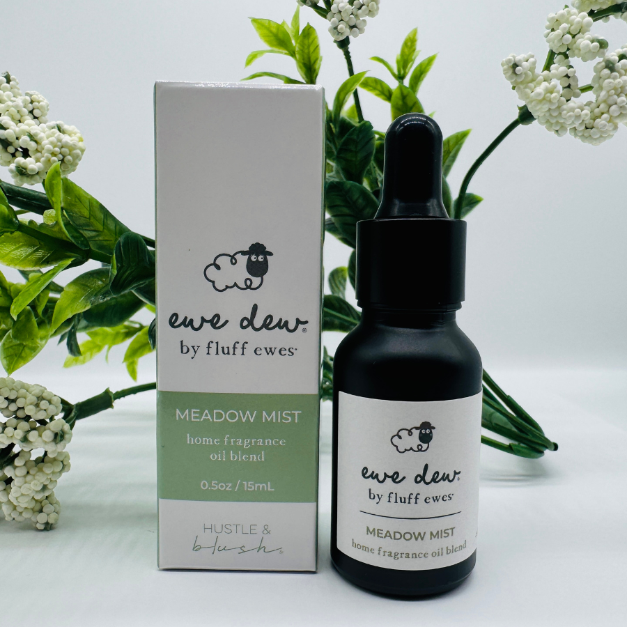 Ewe Dew by Fluff Ewes Meadow Mist Laundry, Home Fragrance + Aromatherapy Essential Oil Blend (0.5 fl oz/15mL) is a light, fresh floral scent for wool dryer balls or oil diffusers, pictured with its box and flowers in the background.