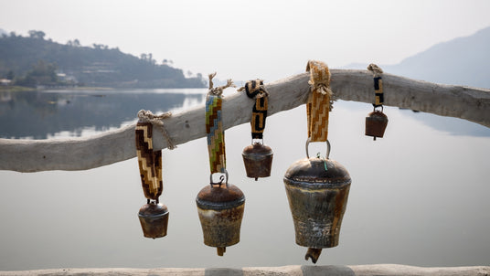 Five rustic cowbells with woven straps hang on a wooden branch beside a calm lake with hills in the background.