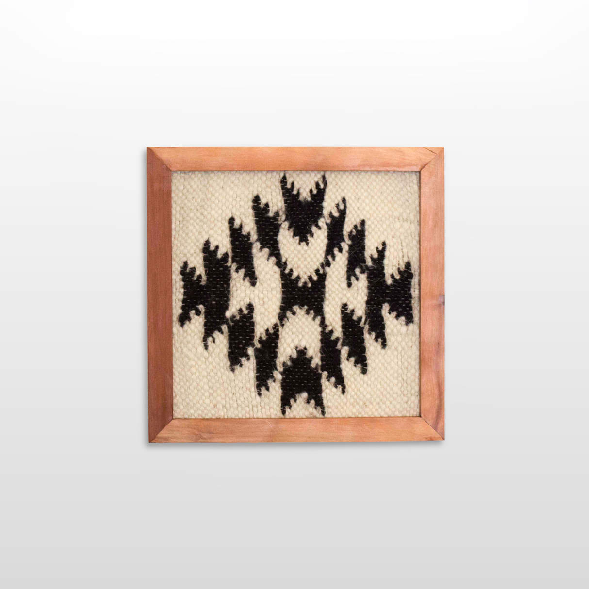 The Vira (Faith) Wool Framed Wall Art, made from undyed sheep’s wool, features a black geometric pattern on beige and is displayed against a plain white wall.