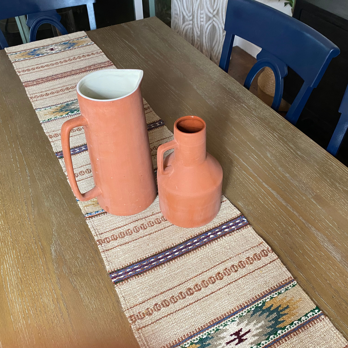 Two orange ceramic pitchers sit on a wool table runner atop a wooden dining table with blue chairs.