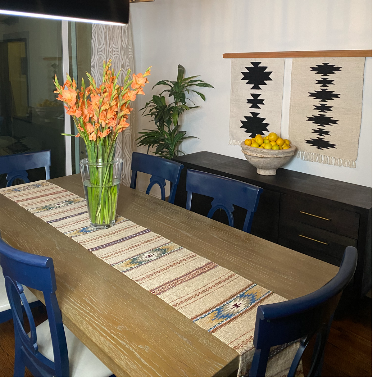 A dining room featuring a wooden table with a wool table runner, blue chairs, orange flowers, and woven wall decor.