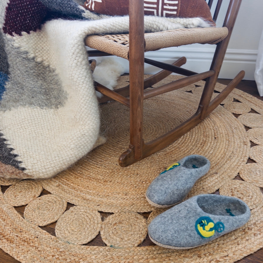 A cozy chair with a knit blanket, Love Ewe Hand-Felted Organic Wool Slippers for Men (Peace-LG), and a woven rug made from organic New Zealand wool on a wooden floor.