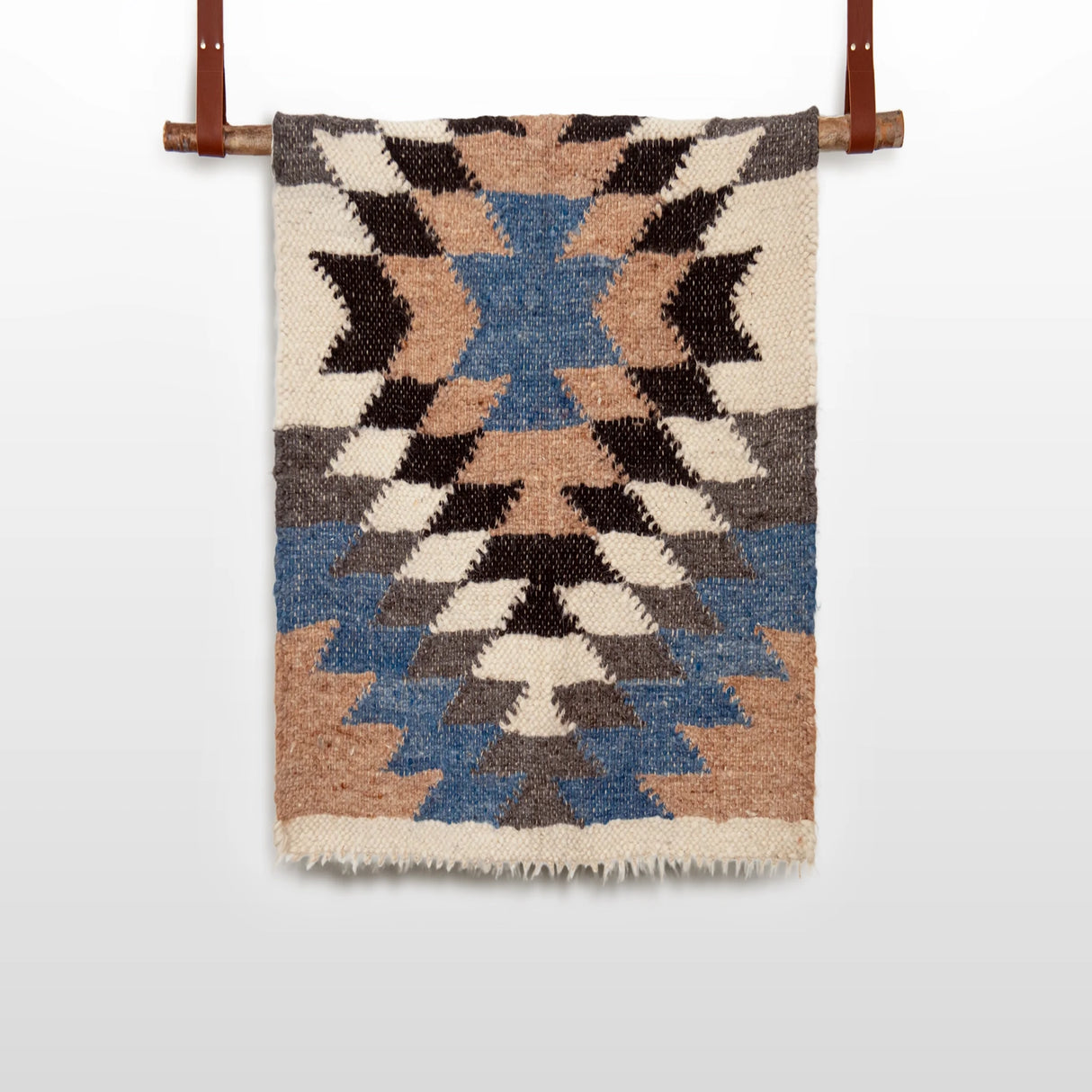 The Zirka (Star) Wool Statement Throw / Baby Blanket, handwoven with blue, brown, black, and cream geometric patterns, hangs on a wooden rod with leather straps.