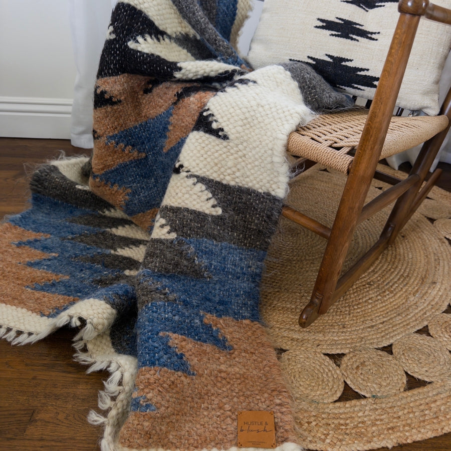 The Zirka (Star) Wool Statement Throw / Baby Blanket with a striking pattern is draped over a wooden chair on a woven rug, set against a white wall.