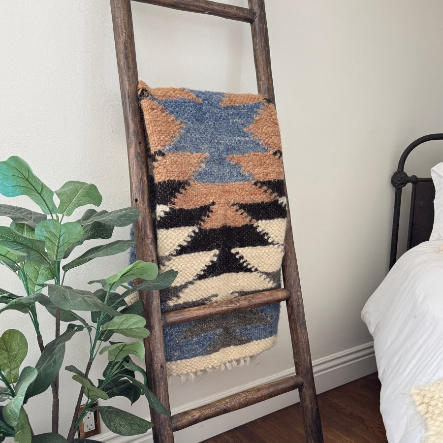 The Zirka (Star) Wool Statement Throw / Baby Blanket hangs on a wooden ladder near a plant and bed in a cozy room.