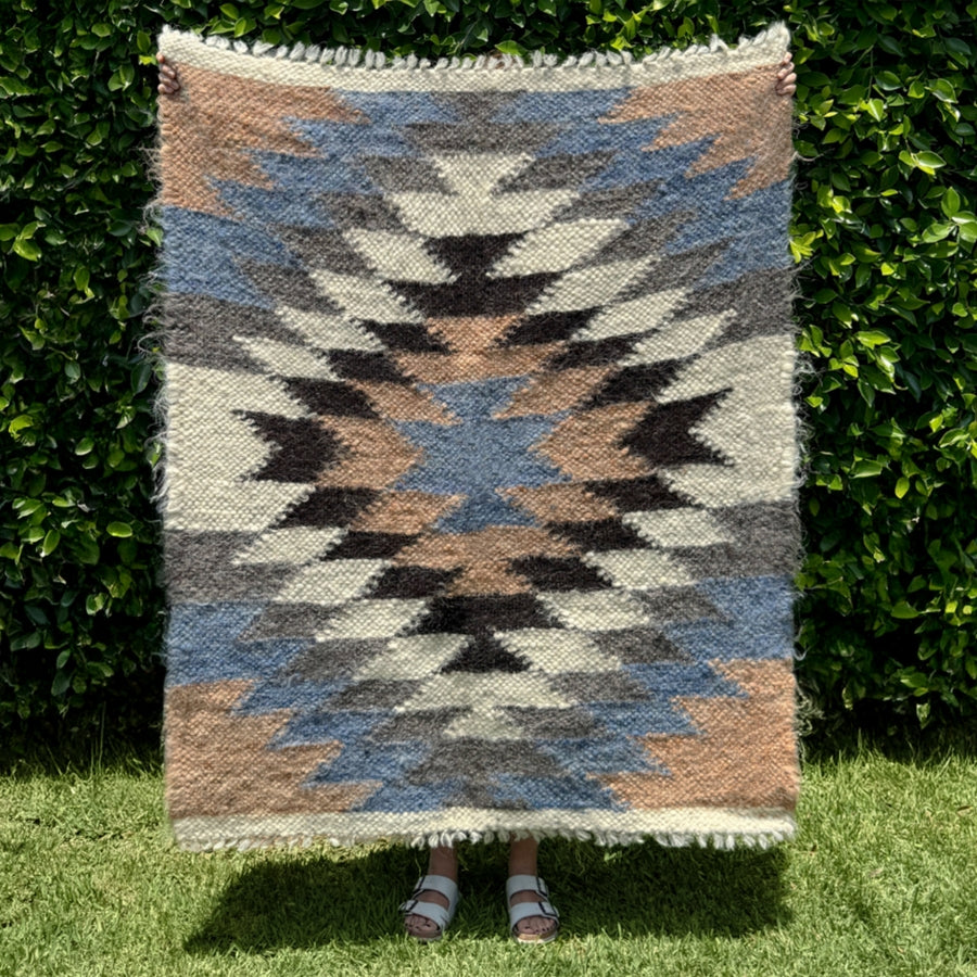 Person holding the Zirka (Star) Wool Statement Throw / Baby Blanket, featuring a geometric pattern and earthy tones, in front of lush green bushes and grass.