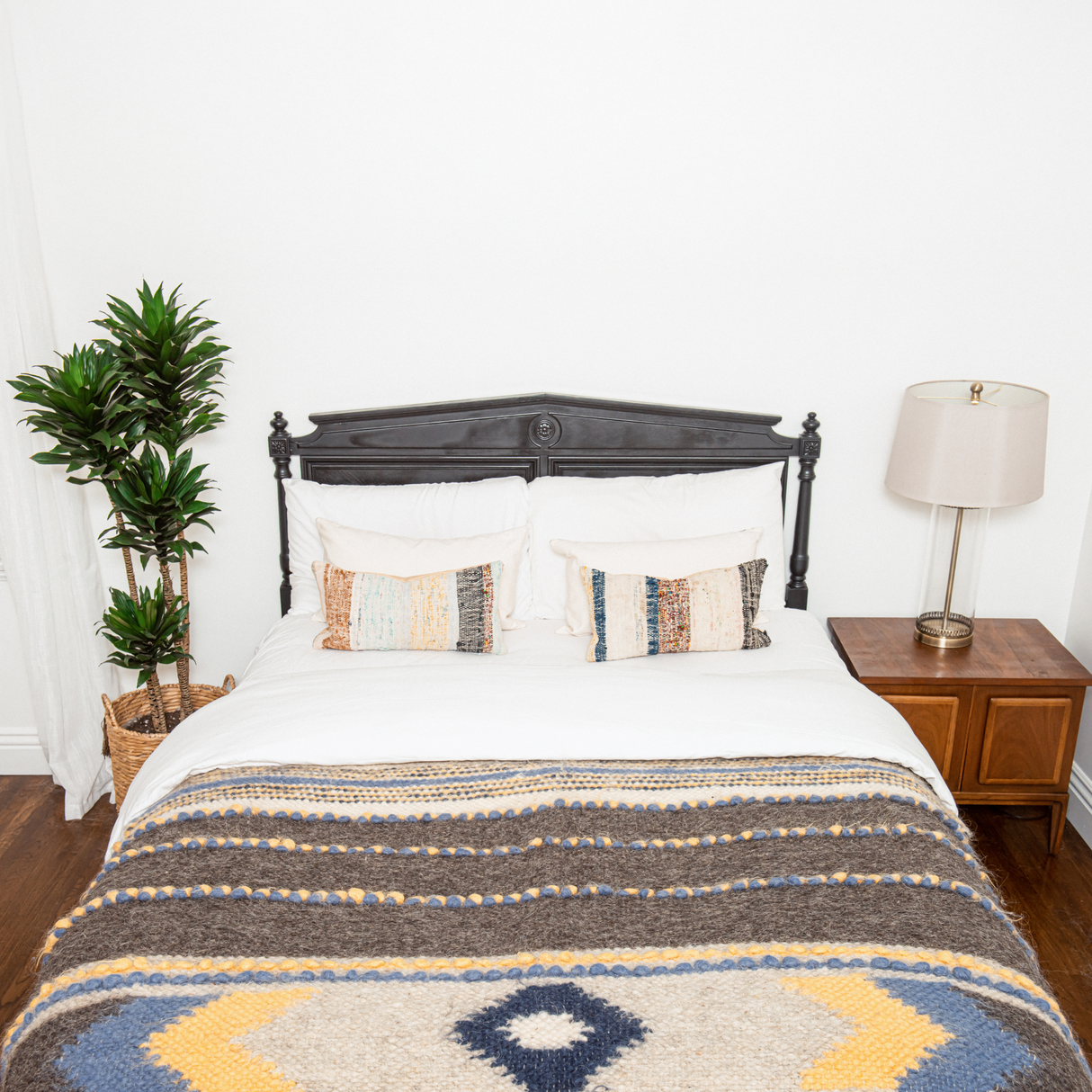 A neatly made bed features patterned pillows and the Zoloty (Golden) Wool Statement Bed Blanket, with a plant and lamp placed on a wooden nightstand.