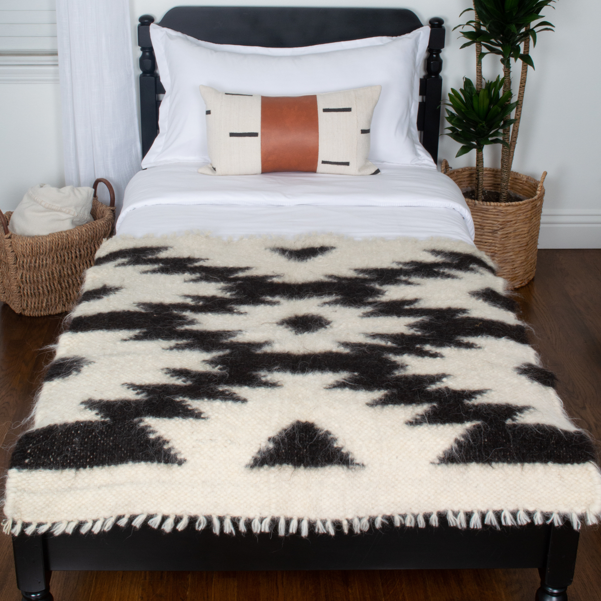 A neatly made bed is styled with the Zytia (Life) Wool Wall Hanging above it, complemented by woven baskets, a plant, and cozy black and white bedding.