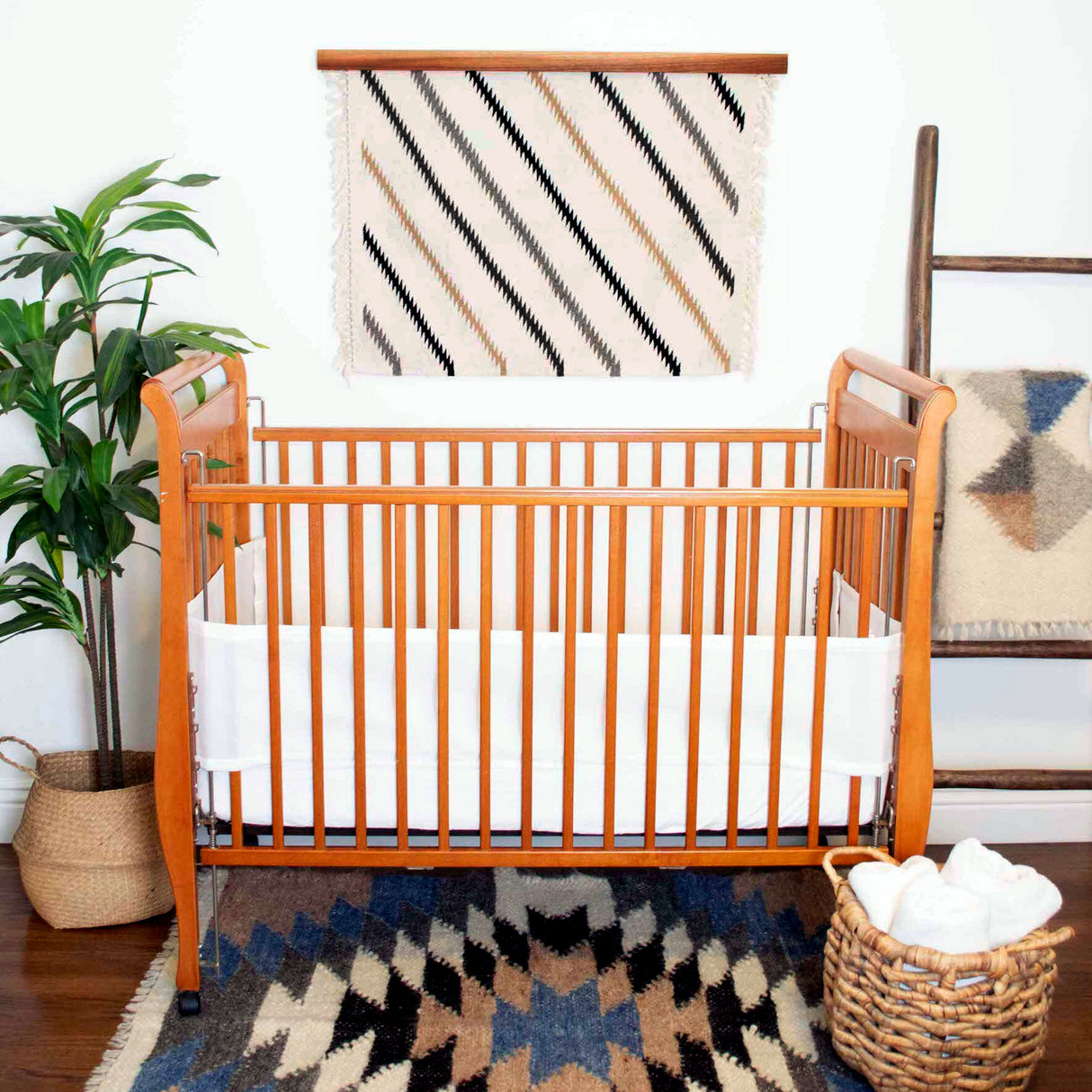 Wooden baby crib with white bedding, handwoven creation blankets, plant, woven basket, and the Promin (Beam) Wool Rug / Wall Hanging add warmth to this cozy nursery.