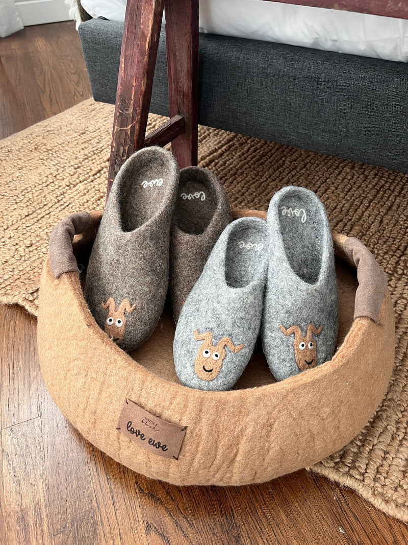 Two pairs of wool slippers with dog faces sit in a round, tan pet bed on a wooden floor.