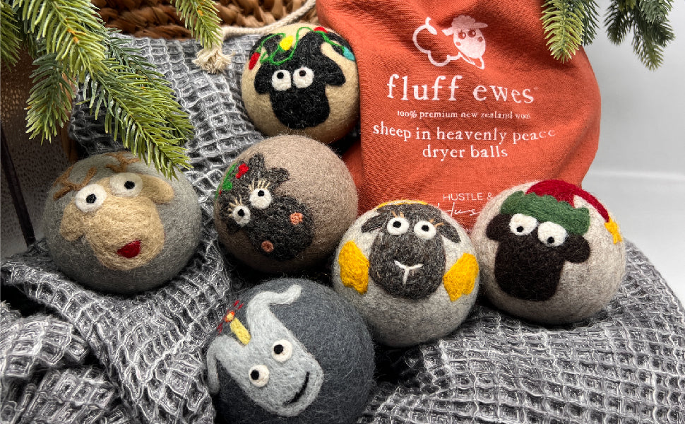 Six Fluff Ewes Premium XL Dryer Balls "Sheep in Heavenly Peace" Holiday Limited Edition (6 Pack), with sheep faces and festive designs, sit on a gray towel beside the orange bag, offering a natural fabric softener alternative.