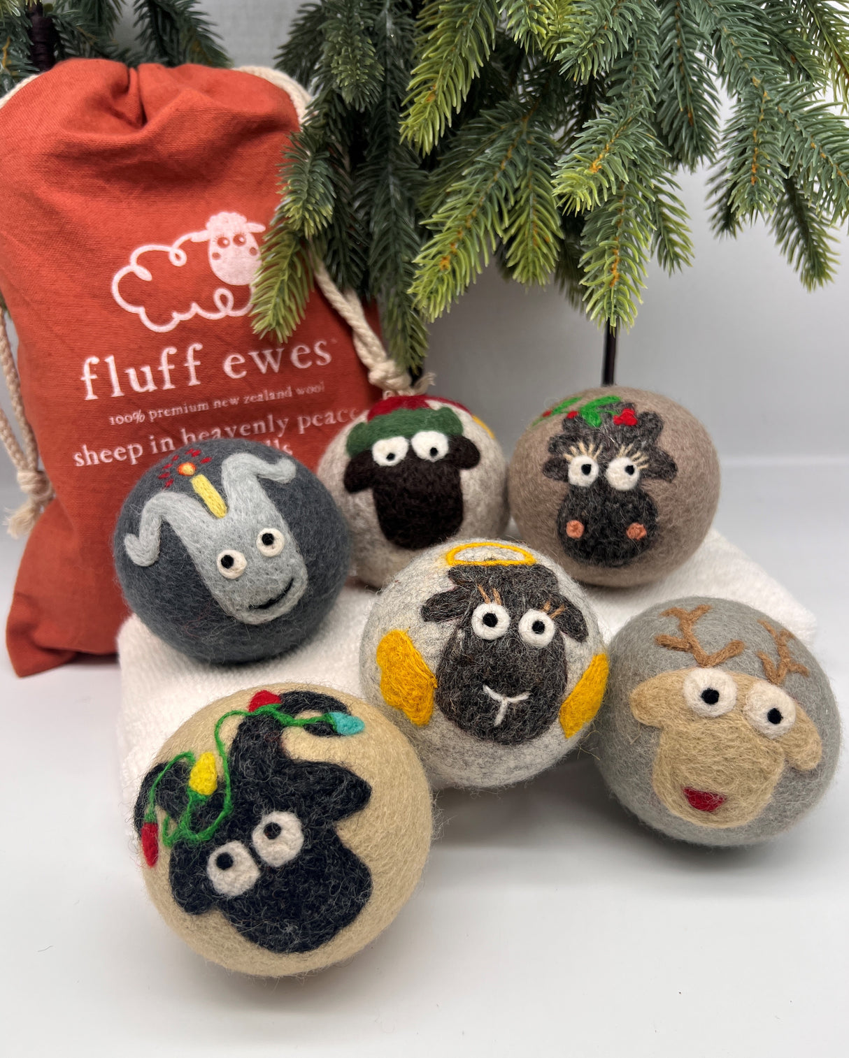 Six Fluff Ewes Premium XL Dryer Balls "Sheep in Heavenly Peace" 6 Pack, Holiday Limited Edition, with cute animal faces and red bag rest under a pine branch—an eco-friendly, natural alternative to traditional fabric softeners.