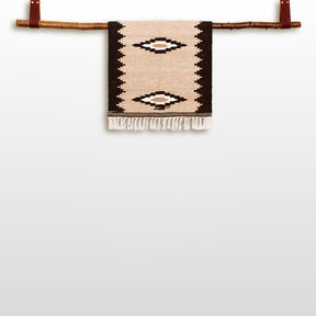 The Heredij (Heritage) Wool Rug, a handmade beige and black geometric wall hanging crafted from natural wool, is displayed on a wooden branch against a white background.