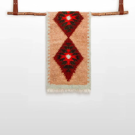 The Mriya (Dream) Wool Bed / Couch Runner - Style B, crafted from Carpathian sheep wool with red diamond patterns, hangs from a wooden rod and highlights Ukrainian artisan textiles against a plain white backdrop.