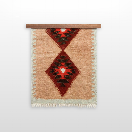 The Mriya (Dream) Wool Bed / Couch Runner - Style B is made by western Ukraine artisans from Carpathian sheep’s wool, featuring two red diamond patterns, fringe, and is presented on a wooden rod.