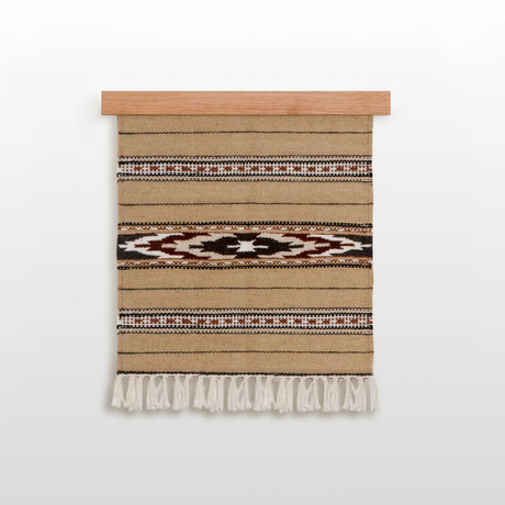 The Teplota (Warmth) Wool Small Rug / Mat is handwoven from organic wool, showcases beige with black, brown, and white patterns, features fringe at the bottom, and is displayed on a wooden rod.